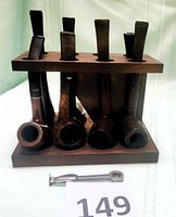 Front view of wooden pipe rack holding seven pipes with different bowls and stems, plus a metal pipe tool in front.