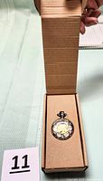 Photo of the pocket watch in original box, cardboard insert with chain folded underneath, watch dial partly visible through transparent section.