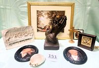 Photo showing all items in the lot arranged on light-colored surface including bronze praying hands, Last Supper plaque, framed Jesus on the Road to Emmaus picture, wooden cross, two oval photos, small framed prayer picture, and Victorian carved cowrie shell.