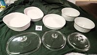 Eight pieces of white CorningWare including four casseroles and three glass lids, and one quiche/tart baking dish on a green cloth.