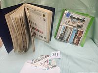Photo of open Ambassador world stamp album displaying various vintage stamps in clear pages, a green binder with old postcards, and an envelope of loose stamps on a table.