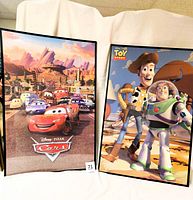 Photo showing two Disney Pixar movie posters for Toy Story and Cars, each in black frames and resting on a white fabric surface.