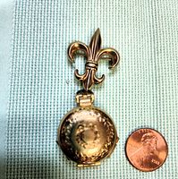 Closed gold tone locket brooch showing fleur-de-lis clasp and hammered circular locket with textured detail side-by-side with penny for size reference.