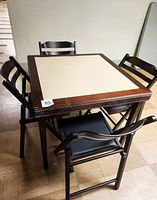 Overall view of the vintage wooden card table with cream inlay top and four matching folding chairs with black faux leather seats arranged around it.