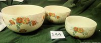 Three cream-colored ceramic mixing bowls nested, showing floral poppy and blue flower designs with gold rims on a green fabric background.