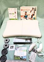 Photo showing Wii Balance Board in front with Wii Fit and Wii Active More Workouts game cases behind it, plus Wii Sports Resort game disc in front. Two white Wii Remote controllers and additional accessories including sensor attachments and balance connector shown.