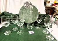 Photo showing 7 clear glass wine glasses with vertical design, crystal decanter with stopper, and vintage round green marble charcuterie board with aluminum decoration on green cloth background.
