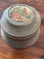 Top view of vintage metal circular powder puff music box showing painted top with woman in a red dress seated and man playing pipe standing in a garden scene.