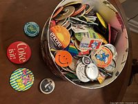 Photo showing multiple vintage buttons in a round container with some laid out including Coca-Cola 'Coke is it!' and a smiley face.