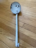 Antique white enamelware slotted spoon with black speckles, perforated bowl, and hooked handle lying flat on a wooden surface.