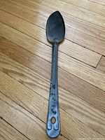 Photo of the entire antique stirring spoon showing metal enamel ware with wear and discoloration, laid on hardwood floor