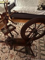 Side view of the antique spinning wheel showing the large wheel, foot pedal, and twisting mechanism on a patterned rug in a room with couch and furniture.