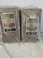 Two sealed packages of Sure Fit short dining chair slipcovers in sand linen color.