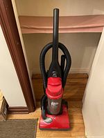 Front view of red Dirt Devil upright vacuum cleaner with flexible hose coiled at back, standing on wooden floor near a beige wall and a brown baseboard.