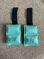 Pair of teal the Cuff ankle and wrist weighted bands with black straps, weight marked 4 lbs (1.81 kgs) each laid on carpet.