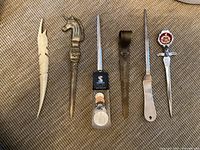 Top-down view of six letter openers showing various designs and materials including carved bone, brass horse head, decorative coins, leather sheath, and red enamel crest.