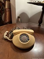 Beige rotary dial telephone showing front view with handset resting on base, coiled handset cord, and attached line cord on wooden table.
