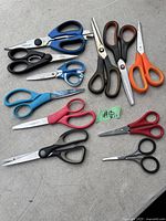Photo of 13 pairs of assorted scissors in various sizes, colors, and handle designs laid out on gray concrete surface.