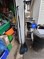 Two Surfstow paddles (AL-20 and AP-25) shown side by side leaning against a wall outdoors among household items.