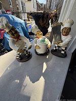 Three vintage chef figurines placed on a ledge outdoors with a background showing a residential area. Figurines are detailed with facial features and utensils such as a cleaver and pots.
