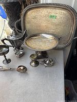All the antique and vintage silver plated items displayed on a surface, including trays, teapot, chalice, salt cellars, candlestick holder, and serving spoon.
