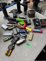 Photo showing a collection of assorted lighters on a table including torch lighters, traditional flip-top, and disposable styles.