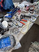 View of assorted tools and hardware on white table, including caulking gun, wire strippers, pliers, hoses, clamps, and hardware in plastic bags.