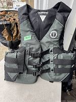Front view of Mustang Survival adult life jacket showing three black adjustable straps and buckles with black and gray nylon fabric, size label L visible.