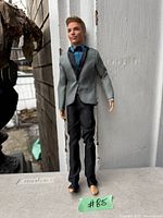 Photo of Barbie Fairytale Groom doll standing outdoors against a white wall background. The doll wears a light grey tuxedo jacket, blue shirt with bow tie, and black pants. Doll is barefoot.