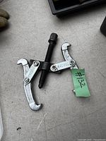 Image showing a metal two-arm bearing puller with central threaded screw and hooked arms placed on a flat surface.