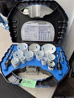 Mastercraft hole saw cutting set inside open black plastic carrying case on outdoor surface. Contains multiple white metal hole saw blades of different diameters, two black mandrels, and two drill bits held in blue foam insert.