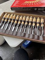 Opened brown leather pouch showing 12 wood carving tools with wooden handles and metal blades in slots.