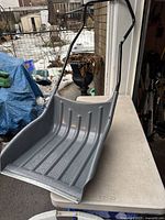 Top-down side view showing the large scoop area of the gray snow sleigh shovel with black metal handles and reinforced metal edge.