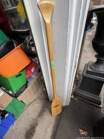 Full length view of the solid wood paddle leaning against a wall showing the entire paddle from handle to blade.