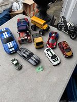 Photo showing ten diecast toy vehicles including Shelby Mustangs, a semi truck with flames, school bus with monster wheels, various cars, and a motorcycle.