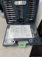 Open plastic case showing assorted taps, dies, handles, and a printed tap drill chart inside the lid