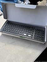 Photo showing a black wireless keyboard with full QWERTY layout inside a white cardboard box, keyboard fully visible.
