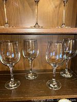 Four clear glass white wine glasses with thin stems standing on a wooden shelf against a wood panel background.