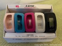 Boxed set displaying one black fitness tracker unit with four interchangeable silicone bands (pink, blue, red, white) and one black band attached to the device.
