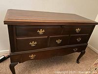 Closed front view of a dark wood chest with five drawers on the front face and brass handles, standing on carved legs.