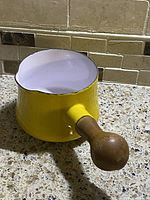 Goldenrod yellow butter warmer pot with white interior enamel and ergonomic wooden handle shown from three angles. Pot displays a spout, rounded body, and flat bottom with signs of use.