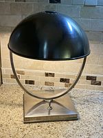 Chrome table lamp with round, perforated metal shade and square base, shown from front angle.