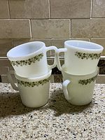 Four vintage Pyrex cups stacked, showing green floral Spring Blossom pattern on white glass, photographed on countertop with tiled backsplash.