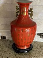 Full view of the red Chinese porcelain vase with gold characters and two ornate gold handles on the neck, placed on a black wooden pedestal