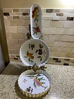 Three white porcelain dishes with detailed fruit motifs displayed vertically on a decorative stand, highlighting scalloped gold rims.