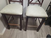 Front view of two brown wooden dining chairs showing beige patterned cushioned seats and wooden legs with stretchers.