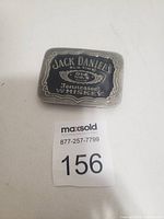 Photo of a metal belt buckle with Jack Daniel's Old No. 7 Tennessee Whiskey logo in black and silver, placed on a white background with MaxSold lot number.