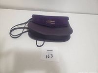 Purple leather purse with thin strap showing front flap and crescent gold-tone metal detail.
