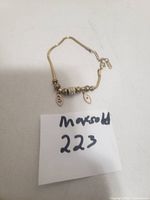 Gold charm bracelet with eye-shaped charms and decorative beads on a thin chain, placed next to a note with item number.