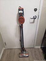 Front full view of an orange and gray stick vacuum cleaner leaning against a white door, showing the vacuum head, handle, and power cord wrapped around the vacuum body.